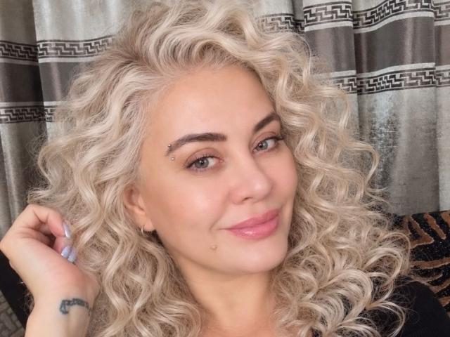 LiyaRose with curly blonde hair, light skin, small nose stud, pink lips, subtle smile, tattoo on left hand, grey eyes, standing in front of grey patterned curtains.