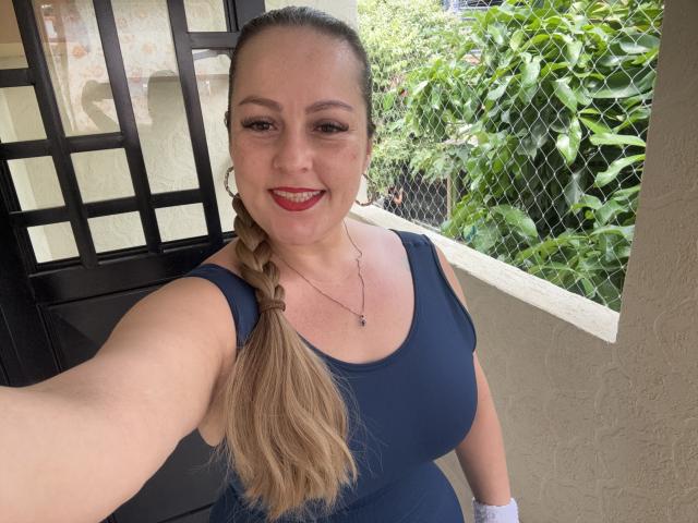 SamanthaGold met een lange blonde vlecht, een blauw tanktopje, rode lipstick, lachend, met grote borsten, een witte horloge, een zwarte deur, een groene, bladerrijke omheining en een bruine muur op een selfie..