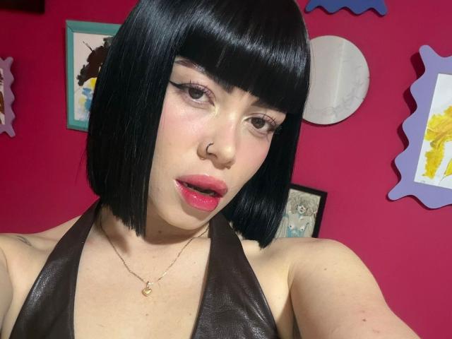 AnaPolaBlairZ with black bob, nose ring, pink lips, brown halter top, gold necklace, looking at camera, pink wall, framed art, and decorative pieces in the background.