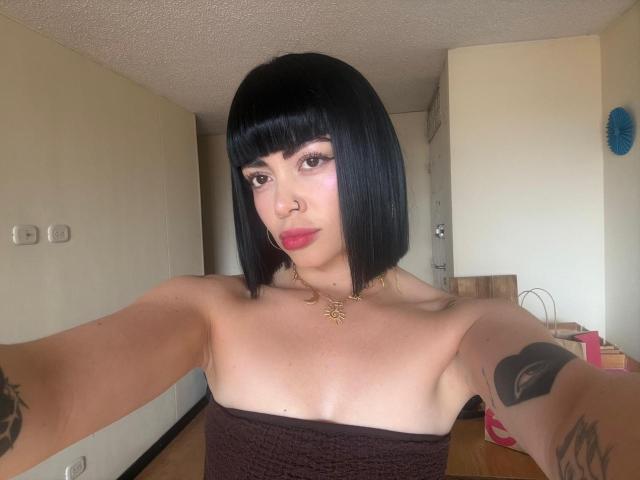 AnaPolaBlairZ, com um corte de cabelo bob preto, piercing no nariz e batom vermelho, vestindo uma blusa sem alças, mostrando seus pequenos seios e tatuagens, tirando uma selfie em um quarto simples..