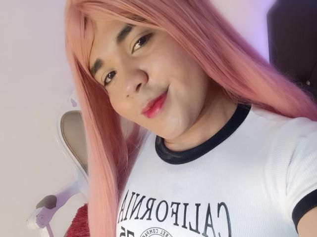 EmilyCavendish with pink hair, brown eyes, and red lipstick, wearing a white shirt with black trim, smiling at the camera, visible boogers on her nose.