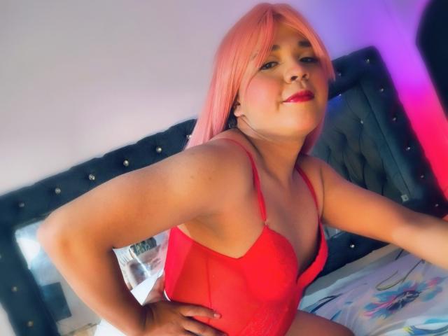 Emily Cavendish mit rosa Haaren, roter Spitzenunterwäsche und rotem Lippenstift, lehnt sich vor, die Hände auf dem Bett, lächelnd und zeigt ihre Brüste. Schlafzimmer mit dunklem Kopfteil..