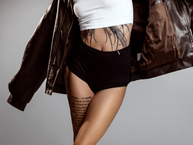 SophieKitty's bare midriff with black panties, white top, and brown leather jacket, showcasing tattoos on her side and lower belly, with a focus on her ass.