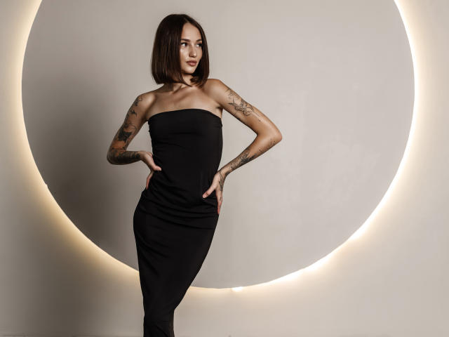 SophieKitty with short brown hair, tattoos on arms, wearing a black strapless dress, hands on hips, standing, looking to the side, in a lit circular background.