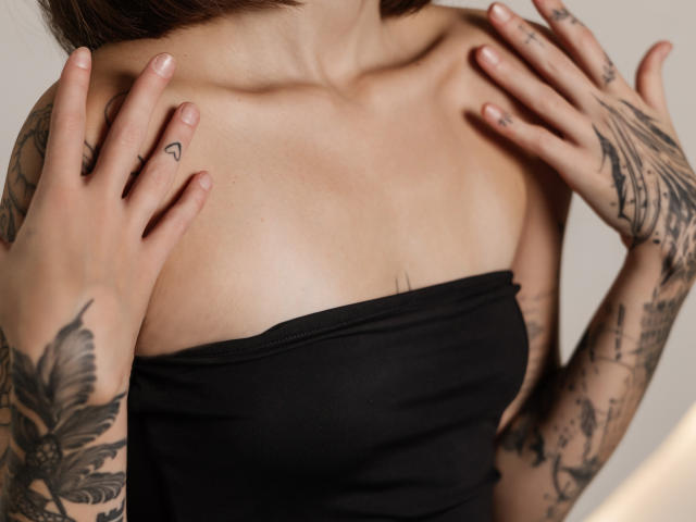 SophieKitty, con tatuaggi sulle mani e sulle braccia, indossa un top nero senza spalline e tocca la clavicola con le dita, mostrando un piccolo tatuaggio a forma di cuore. Il suo seno è visibile..