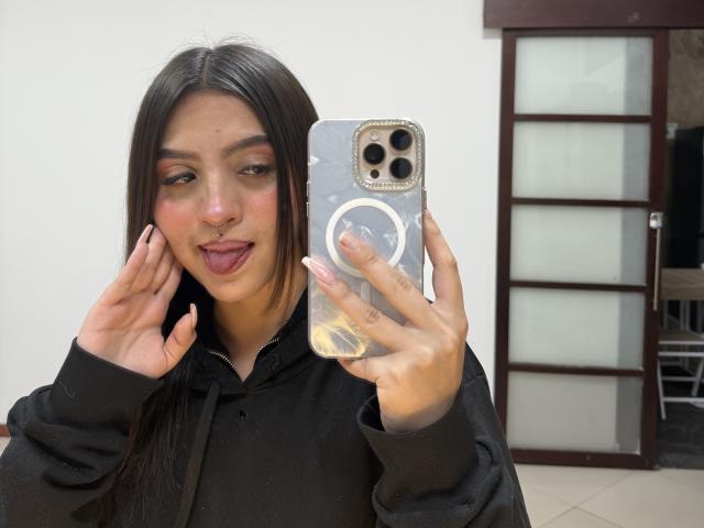LunnaBlossom, com cabelos longos e escuros, moletom preto e piercing no nariz, com a língua de fora, segurando um telefone com um suporte de anel, com os dedos tocando as bochechas. Um iPhone na mão, com uma porta de vidro refletiva ao fundo..