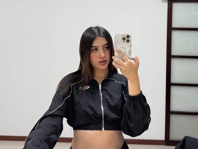LunnaBlossom with dark brown hair, wearing a black zip-up sports jacket, taking a selfie with an iPhone, showing her midriff and slightly open jacket. Her lips are slightly parted.