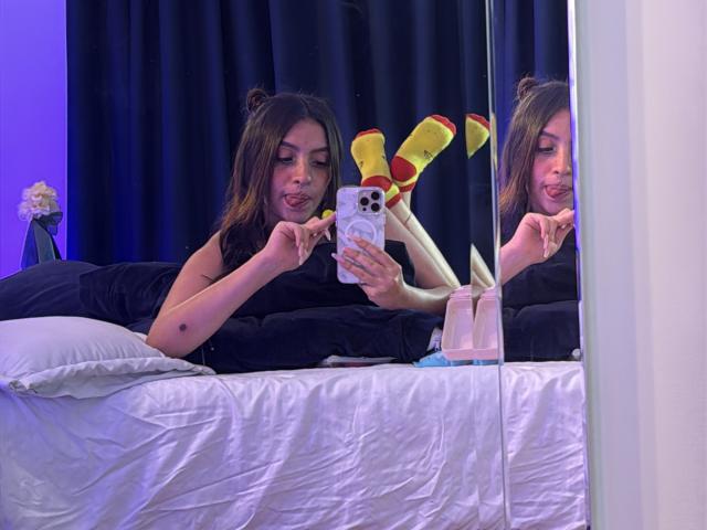 LunnaBlossom with brown hair, lying on a bed, taking a selfie, wearing yellow socks with red dots, black shirt, smiling, mirror reflection, white pillow, black bedsheet.