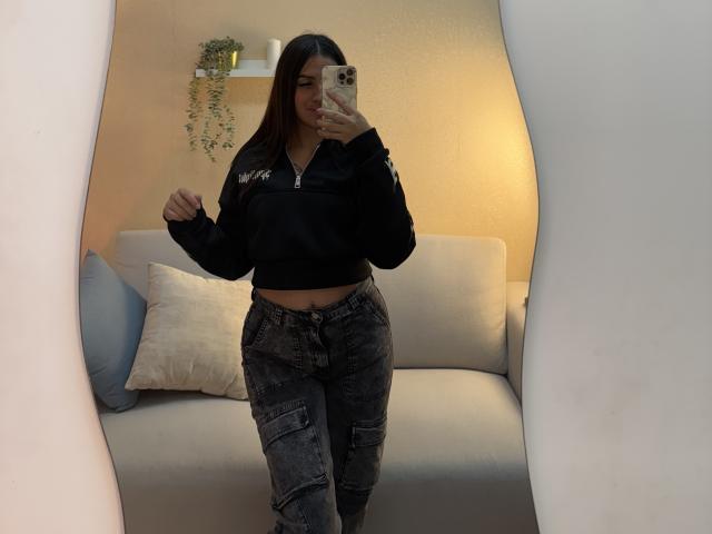 LunnaBlossom with long brown hair, wearing a black crop top and gray cargo pants, taking a mirror selfie, showing her flat midriff and small ass, in a cozy room.