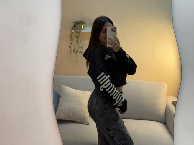 LunnaBlossom with long brown hair, wearing a black hoodie and striped sleeves, gray cargo pants, taking a mirror selfie, showing her curvy ass and small tits.