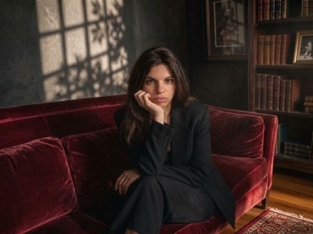 Cicely Dence, com longos cabelos escuros, está sentada em um sofá de veludo vermelho, vestindo um terno preto, com a cabeça apoiada na mão, com uma expressão séria, e sombras da janela em seu rosto..