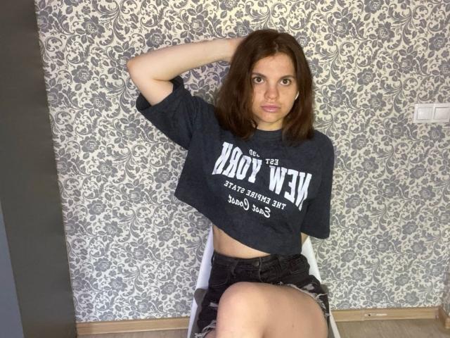 CicelyDence with brown hair, wearing a dark gray "XOXYXOXYXOXY" t-shirt and black shorts, sitting on a white chair, hand in hair, looking at camera, midriff exposed.