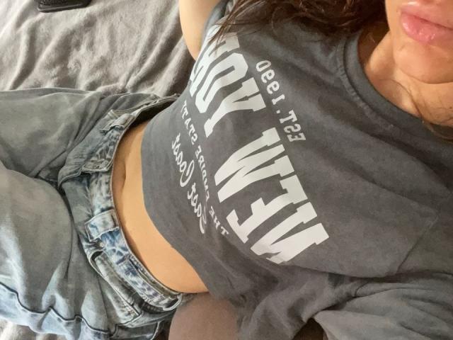CicelyDence veste uma camiseta cinza com texto branco, que mostra seus seios, e está usando jeans azuis com o umbigo à mostra. Seus lábios estão ligeiramente entreabertos..