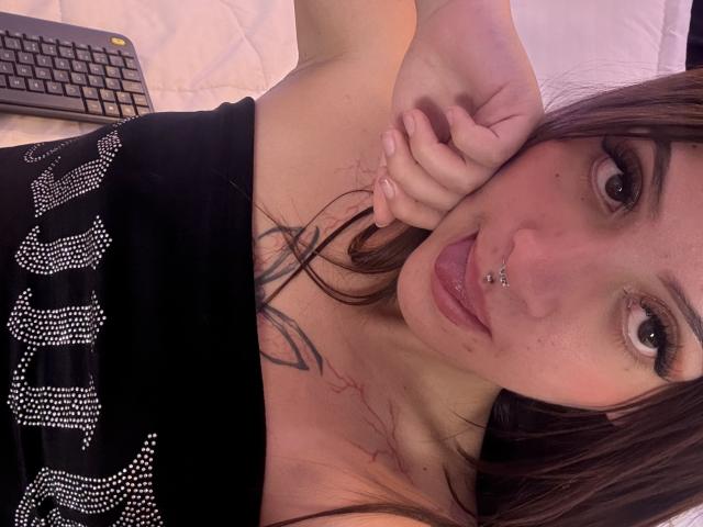 LunnaBlossom, com a língua de fora, piercing no nariz e cabelo castanho comprido, deitada na cama, vestindo uma blusa preta com letras brilhantes, com tatuagens no peito, um laptop ao fundo, e a mão perto do rosto..