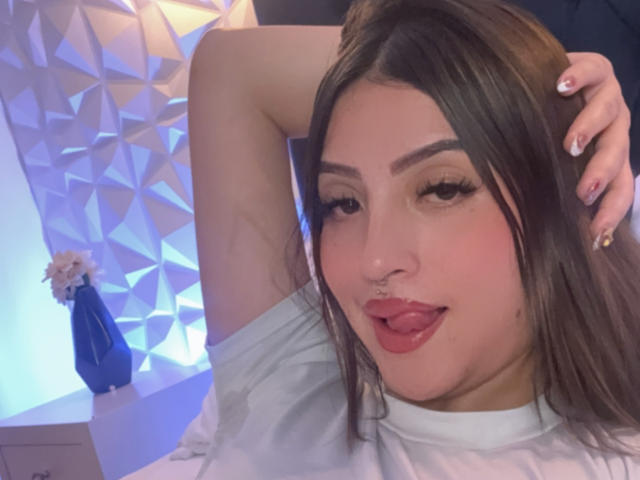 LunnaBlossom with brown hair, nose ring, and tongue out, sticking up her arm, wearing a white shirt, has long eyelashes and painted nails, looking at the camera.