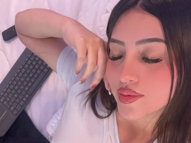 LunnaBlossom with pouty lips, nose ring, and long eyelashes, lying on a bed with a laptop, her hand near her ear, wearing a white shirt.