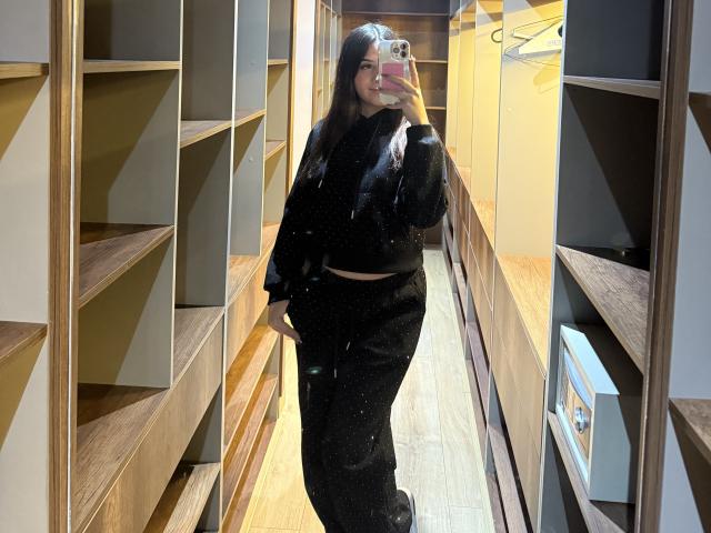 LunnaBlossom with long dark hair, wearing a black fuzzy crop top and black pants, taking a mirror selfie in a wooden room, holding a pink phone.