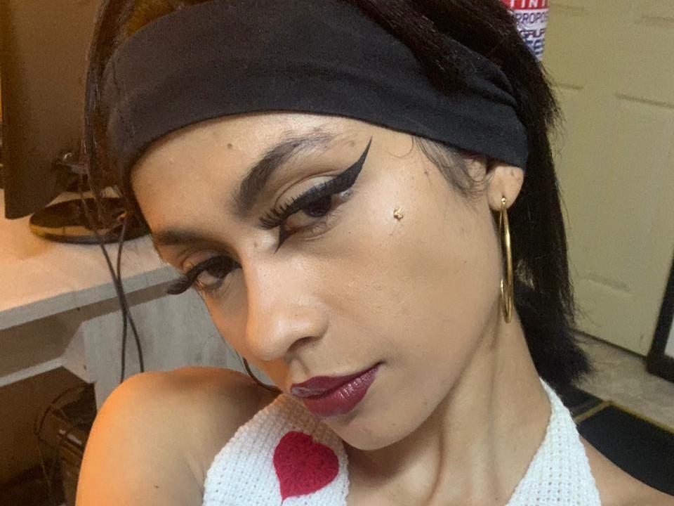 HeidyRusso with dark hair in a black headband, wearing bold black eyeliner, gold earrings, and red lipstick, in a white top with a red heart. Her skin is tan.