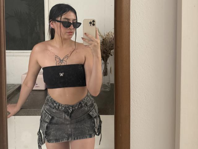 LunnaBlossom with black tube top showing small tits, butterfly tattoo, denim skirt, black sunglasses, holding iPhone, mirror selfie, dark hair, pale skin, tattoos, standing indoors.