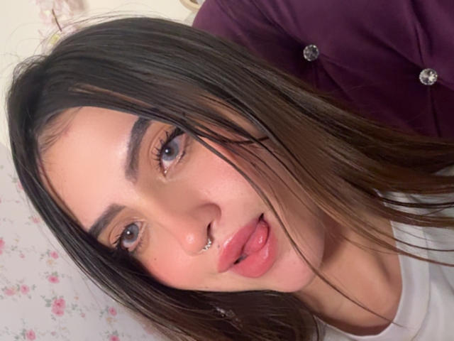 LunnaBlossom has long brown hair, a nose piercing, and is sticking her tongue out with pink lips. She has blue eyes and a white top.