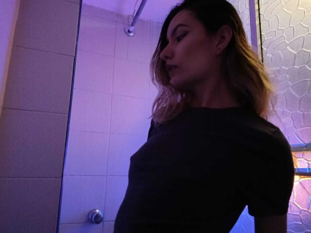 AlanisBlanc, with side profile, wearing a black shirt, visible small tits, in a dimly lit bathroom, blonde hair, purple light, looking to the side, slightly pouty expression.