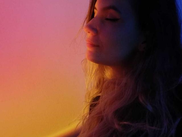 AlanisBlanc with closed eyes, soft smile, and wavy pinkish-brown hair, illuminated by colorful gradient light, showing warm and cool tones on her face.