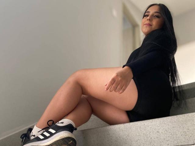 StefanyDelly with long black hair, black long-sleeve top, black shorts, and black Adidas sneakers with white stripes, sitting on gray stairs, showing her ass and legs.