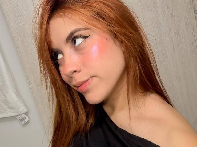 OriRivass tiene el pelo largo y rojo, una ligera sonrisa y mejillas rosadas. Está mirando hacia un lado, con delineador de ojos negro y un piercing en la nariz. Lleva una camiseta negra..