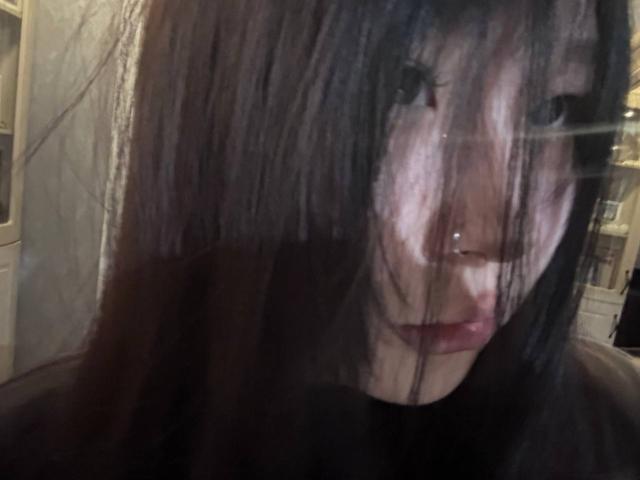 JungMcgranor with messy black hair, nose piercing, and slightly parted red lips, looking through a blurry glass surface, showing subtle reflection and soft light on her face.