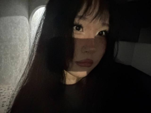 JungMcgranor with long black hair, brown eyes, and a nose ring, looking into the camera, shadowed face, red lips, dark shirt, soft light on the left.