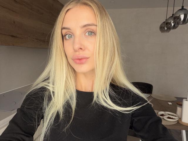 Chanel Paskow a les cheveux blonds, les yeux bleus et les lèvres roses, elle porte un haut noir et a une expression neutre, dans une cuisine moderne avec des murs en bois et des luminaires suspendus..