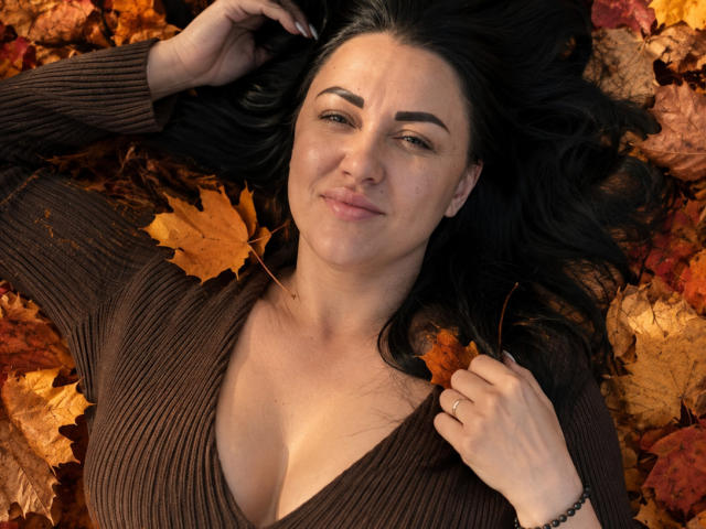 CorneliaRosie is lying on autumn leaves, wearing a brown ribbed top with a deep cleavage, black hair, and a slight smile, holding her hair with one hand.