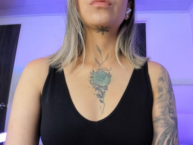 KathyM with blonde hair, black tank top, green rose tattoo on chest, bee tattoo on neck, and tattooed arm, showing her tits and upper body.