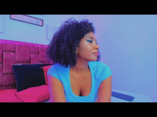 MeganWlliams with curly black hair, wearing a blue top, showing her tits, sitting on a bed with pink and black pillows, looking to the side with a pouty expression.