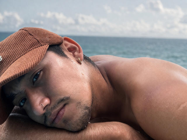 Shirtless RyanTakeda with tan skin, brown cap, and gold earring, laying down with head on arm, blue eyes, and slight stubble, near a calm ocean.