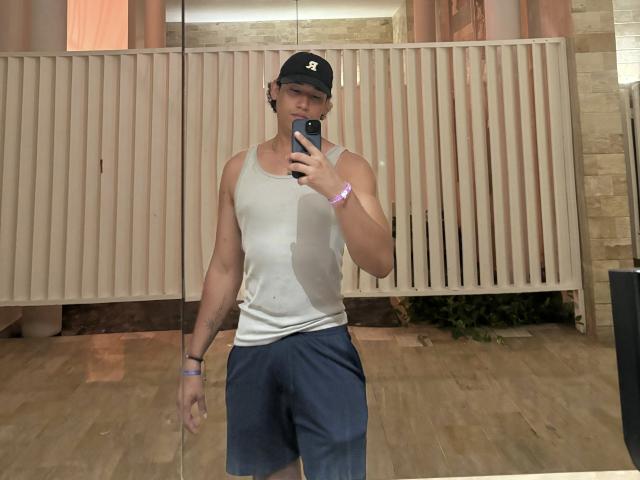 RyanTakeda with a slim, muscular build, wearing a white tank top, blue pants, black cap, and purple wristband, taking a mirror selfie, showing a slightly visible penis bulge.