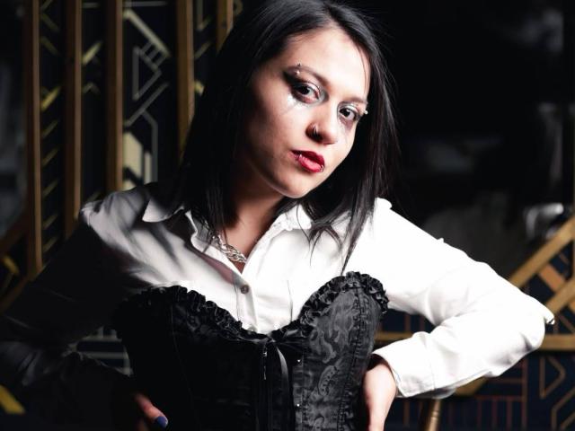 GoddessLuxliv with black hair, white shirt, and black corset, hands on hips, red lipstick, eye makeup, and a nose ring, looking confident. Boobs are accentuated by the corset.
