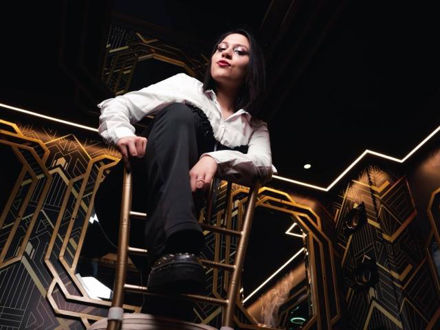 GoddessLuxliv with pale skin, black hair, red lipstick, white shirt, black pants, and black shoes, standing on stairs, looking down, low angle shot, confident, powerful, dominant.