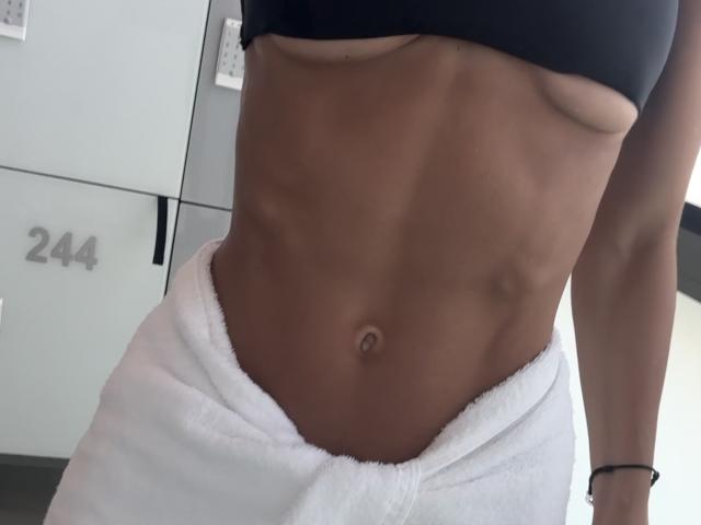 Fridaa's toned, brown-skinned abs with visible muscle definition, wearing a black sports bra and white towel around her waist, a black bracelet on her right wrist.