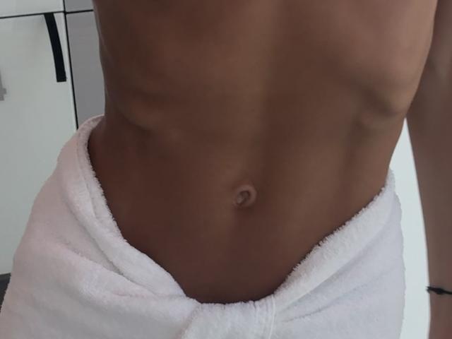 Fridaa's toned, brown-skinned abs with a visible navel, wearing a white towel wrapped around her lower waist. Her slim, muscular body is partially shown.