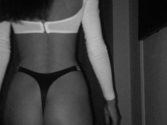 Black and white photo of Fridaa's back showing her bare ass in a thong, wearing a white long-sleeve top with a zipper, and her hands down by her sides.