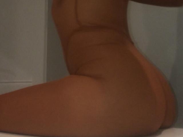 Fridaa's brown, curvy ass is in the foreground, with her side showing in a slightly turned, kneeling position. Her skin is smooth and she's wearing sheer, dark underwear.