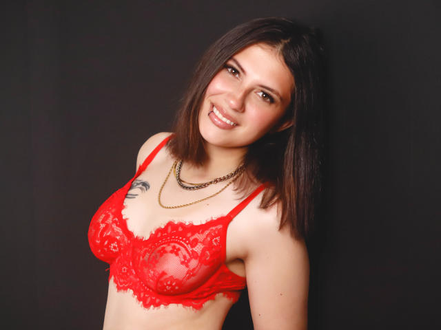 MaryCruzz with red lace bra, smiling, brown hair, necklace, small tattoo on left chest, medium tits, fair skin, black background, standing, looking at camera.