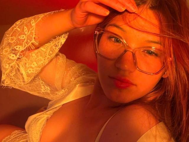 ElodieGardner with red-orange lighting, wearing transparent glasses, white lace top, and pink lipstick, touching her hair, showing her tits, and a soft, confident expression.