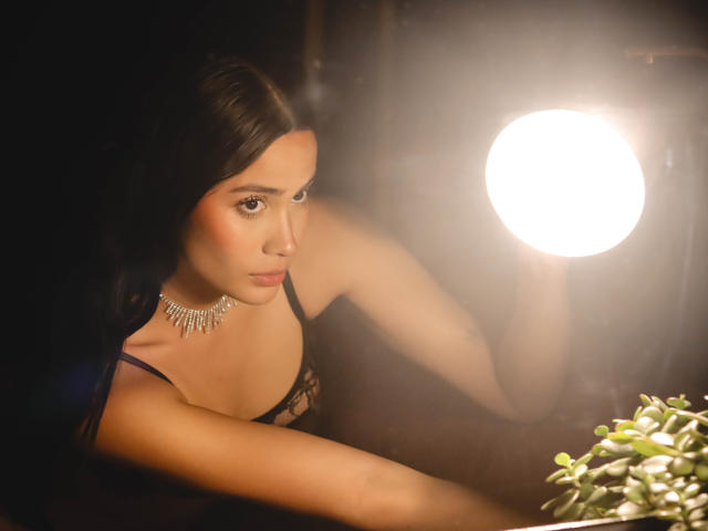 JennTylor with dark hair, wearing a black lace top and silver necklace, looking at a bright light, her arm on a plant, soft light on her face.