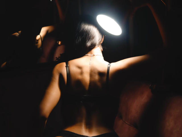 JennTylor's back is to the camera, showing off her toned ass and black bra straps. She's under a bright ring light, with dark hair and a necklace.