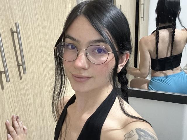 EvaaFarix with glasses, black braided hair, black halter top, showing ass in mirror, blue shorts, tattoo on right shoulder, wood cabinet, reflection, light skin, slight smile.