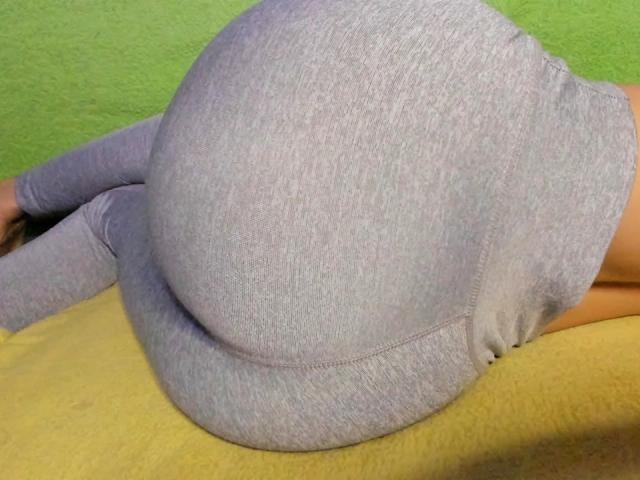 De grote, ronde billen van HarlyMia, in strakke, lichtgrijze, geribbelde leggings, liggen op een gele ondergrond met een groene achtergrond..