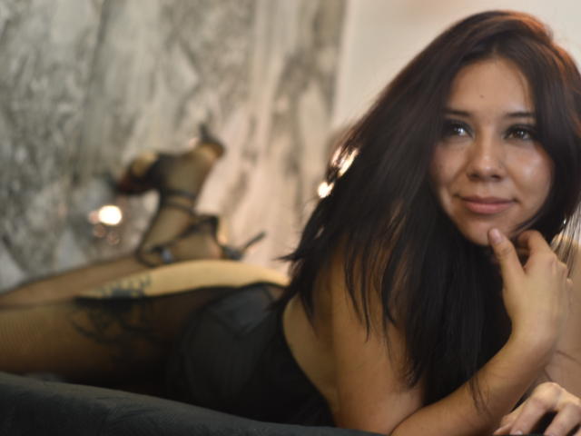 Agatta Adams mit dunkelbraunen Haaren, liegt auf einem schwarzen Sofa, trägt schwarze Dessous, Strumpfhosen und High Heels, stützt ihr Kinn mit der Hand ab und lächelt sanft..