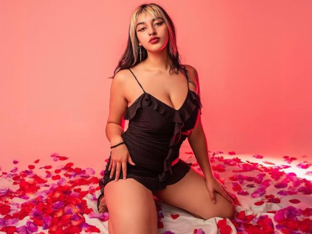CatBleu with blonde and black hair, wearing a black ruffled dress, kneeling on a white bed covered in red and pink petals, looking at the camera with a serious expression.